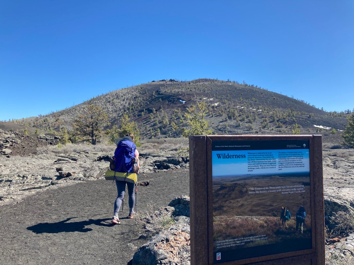 A backpacker hikes on a wide trail with a hill in the background and a wilderness sign in the foreground.