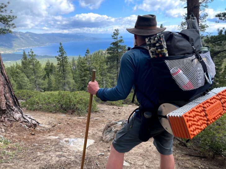 a man stands on an overlook looking out at a lake. The pack he's wearing has a sleeve for holding water bottles sewn into the hipbelt. The water bottle sits at his hip.