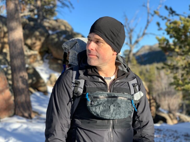 A man stands facing the camera and wearing a backpack with an extra pouch at the chest.