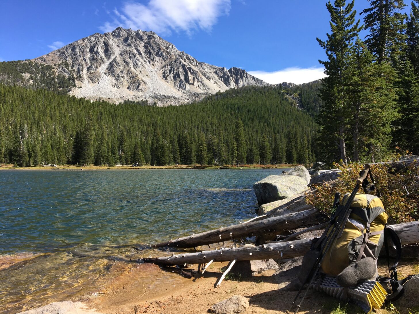 a yellow backpack sits on the edge of a lake with a mountain in the background. Various gear is strapped to the outside of the pack.