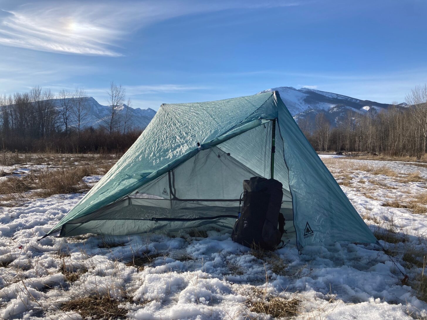 A tent and background are pitched on top of snow with mountains in the background.