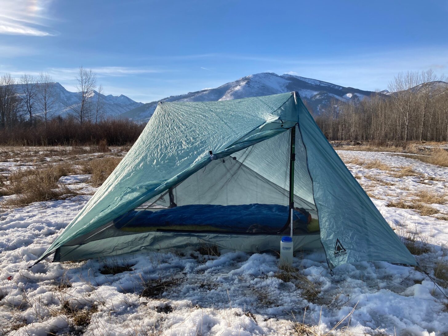 A tent and background are pitched on top of snow with mountains in the background.