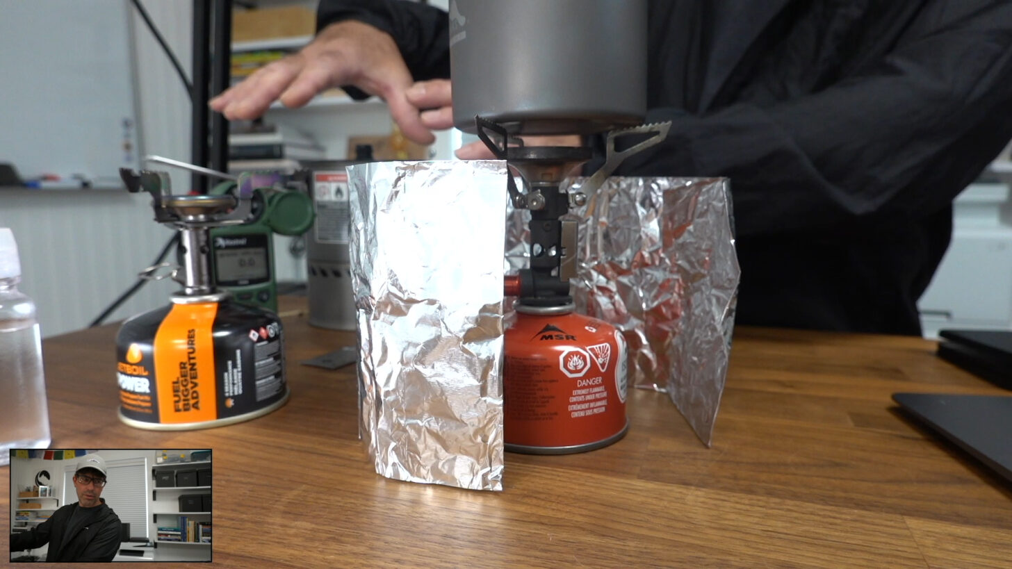 a hand gesturing to a stove surrounded by an aluminum foil windscreen.