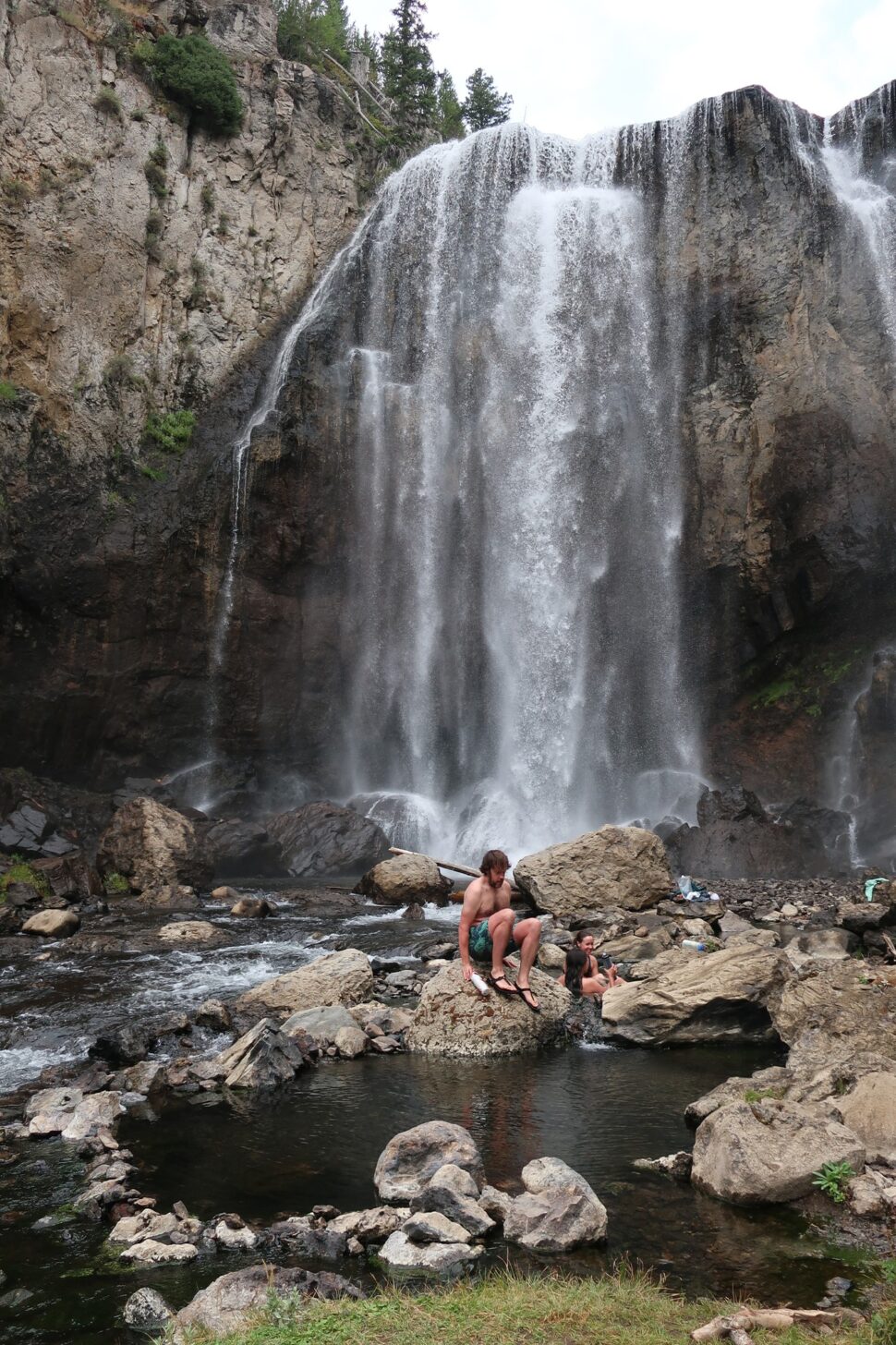 A waterfall is in the background and a hot spring and people are in the foreground.